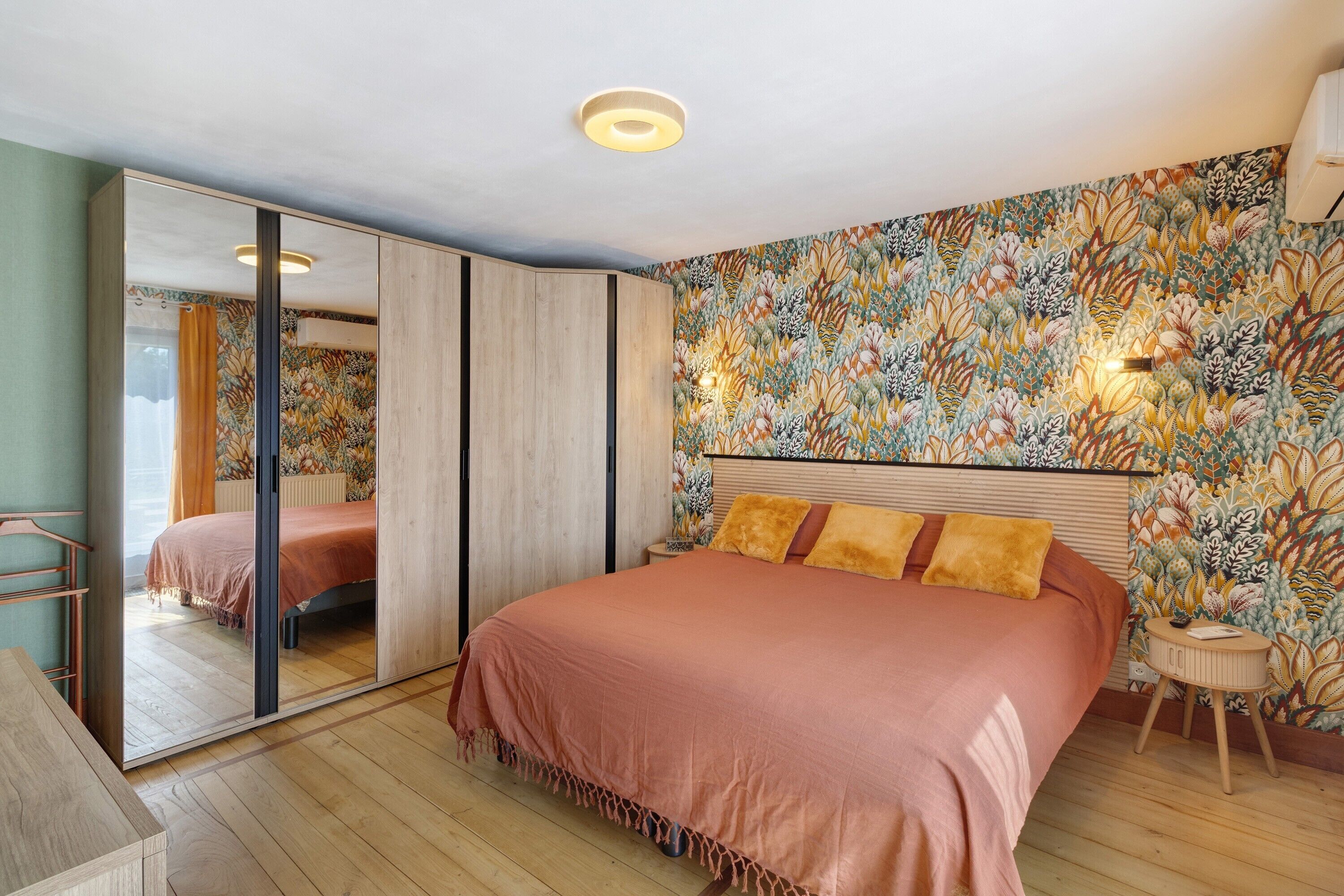 Un dormitorio con una cama grande, un espejo, un armario, una mesita pequeña y una pared con estampado floral.