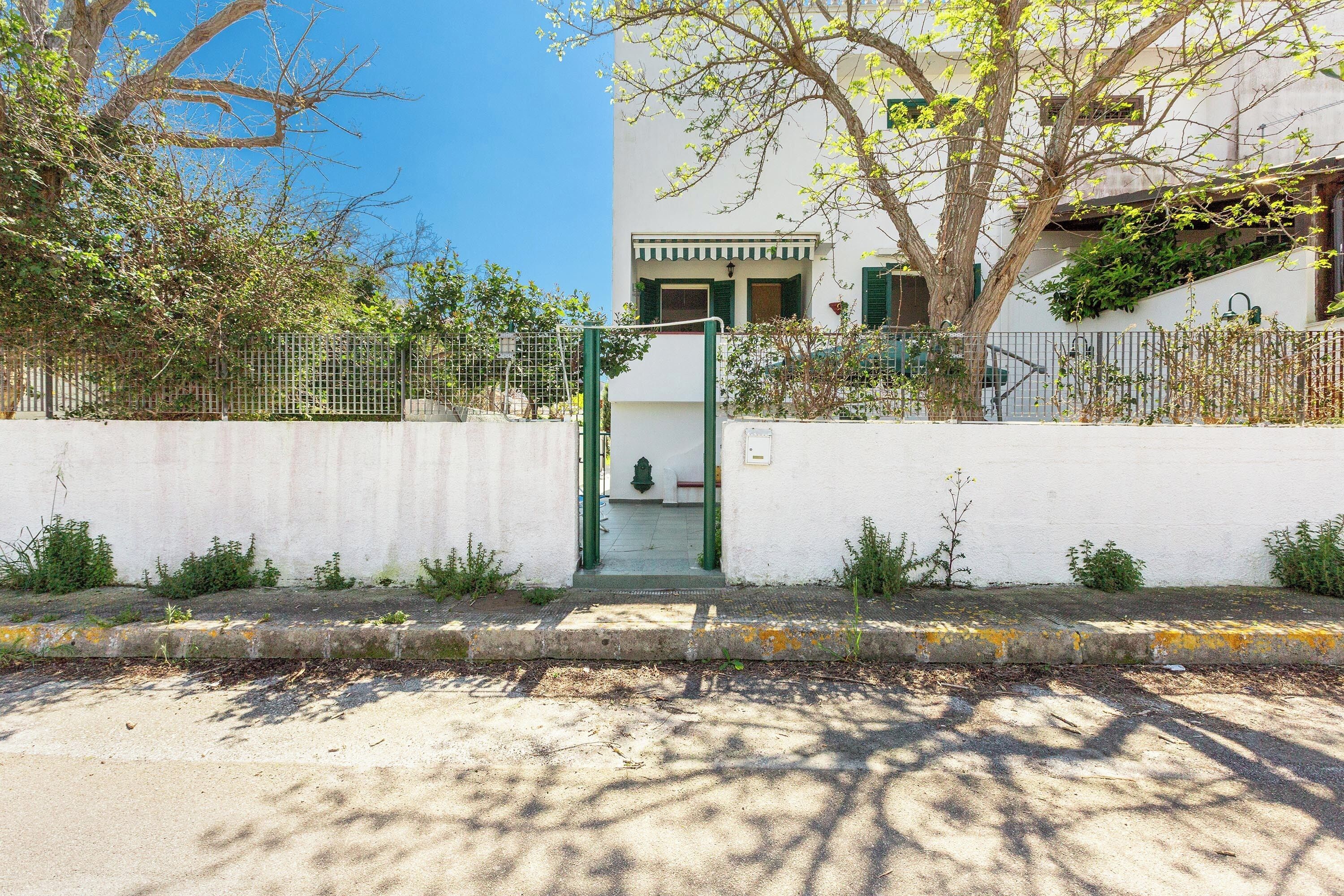A white building with a green gate, a concrete sidewalk, and trees.