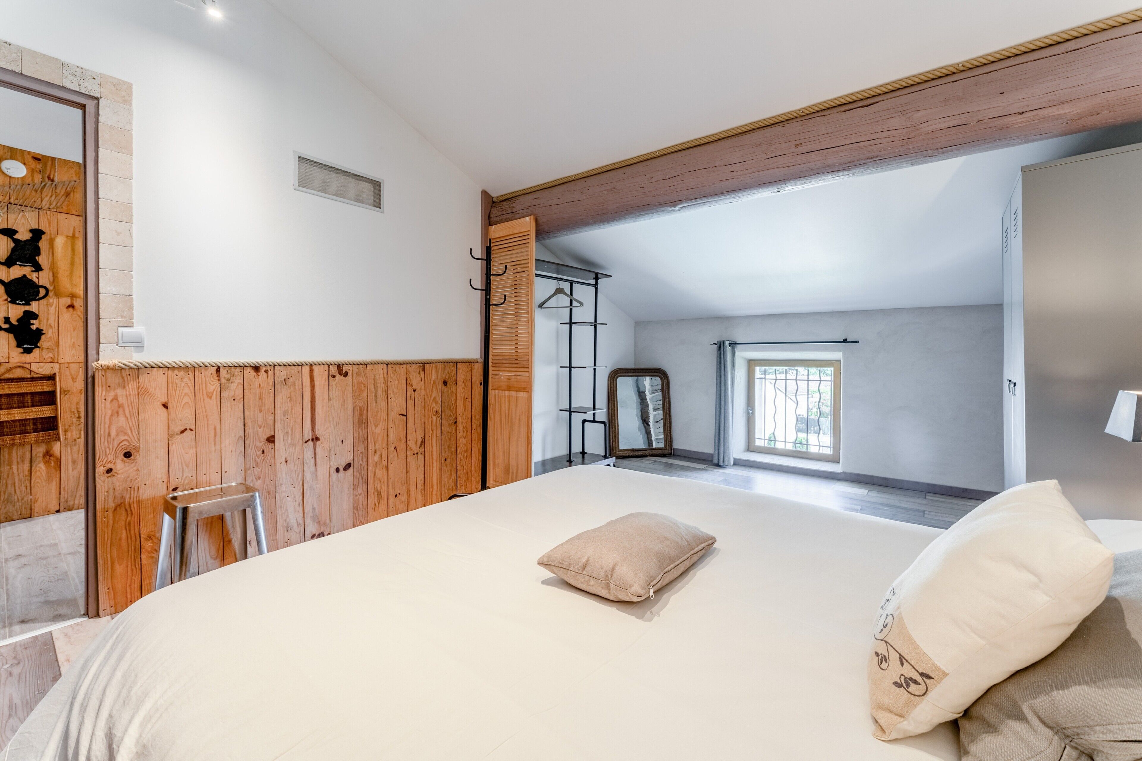 A bedroom with a wooden wall, a bed with pillows, a hanging coat rack, and a window with a view.