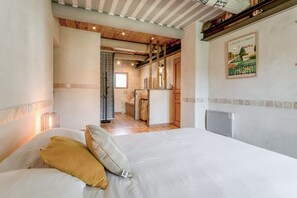 Un dormitorio con cama, almohadas, una mesita de noche, techo de madera, una cocina al fondo y un cuadro en la pared.