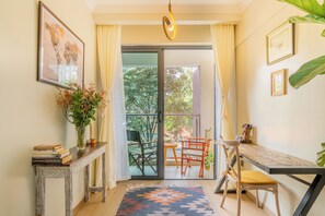 Ein Zimmer mit einer Glastür zum Balkon, einem kleinen Tisch mit Stühlen, einem Blumentopf und einem gerahmten Bild an der Wand.