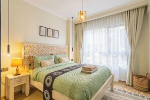 Ein Schlafzimmer mit Bett, Nachttisch, Lampe, Pendelleuchte, Fenster mit Vorhängen und einem gewebten Korb.