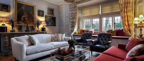 A living room with a white sofa, red armchair, and a black chair. There are books on a glass coffee table, a chandelier, and a window with curtains.