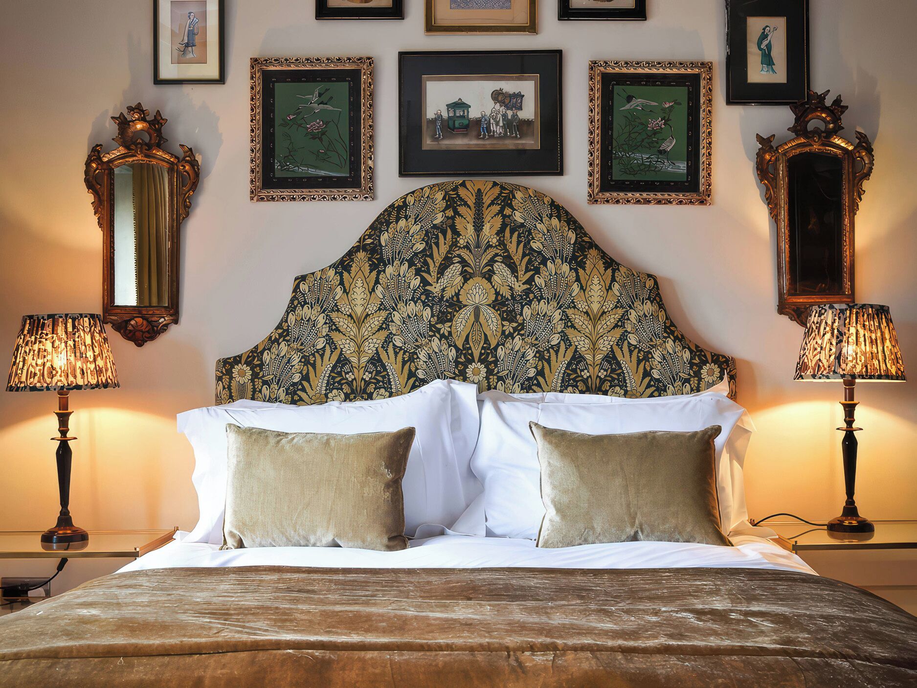 A bedroom with a large bed, two bedside lamps, a headboard with a floral pattern, and framed artwork on the wall.