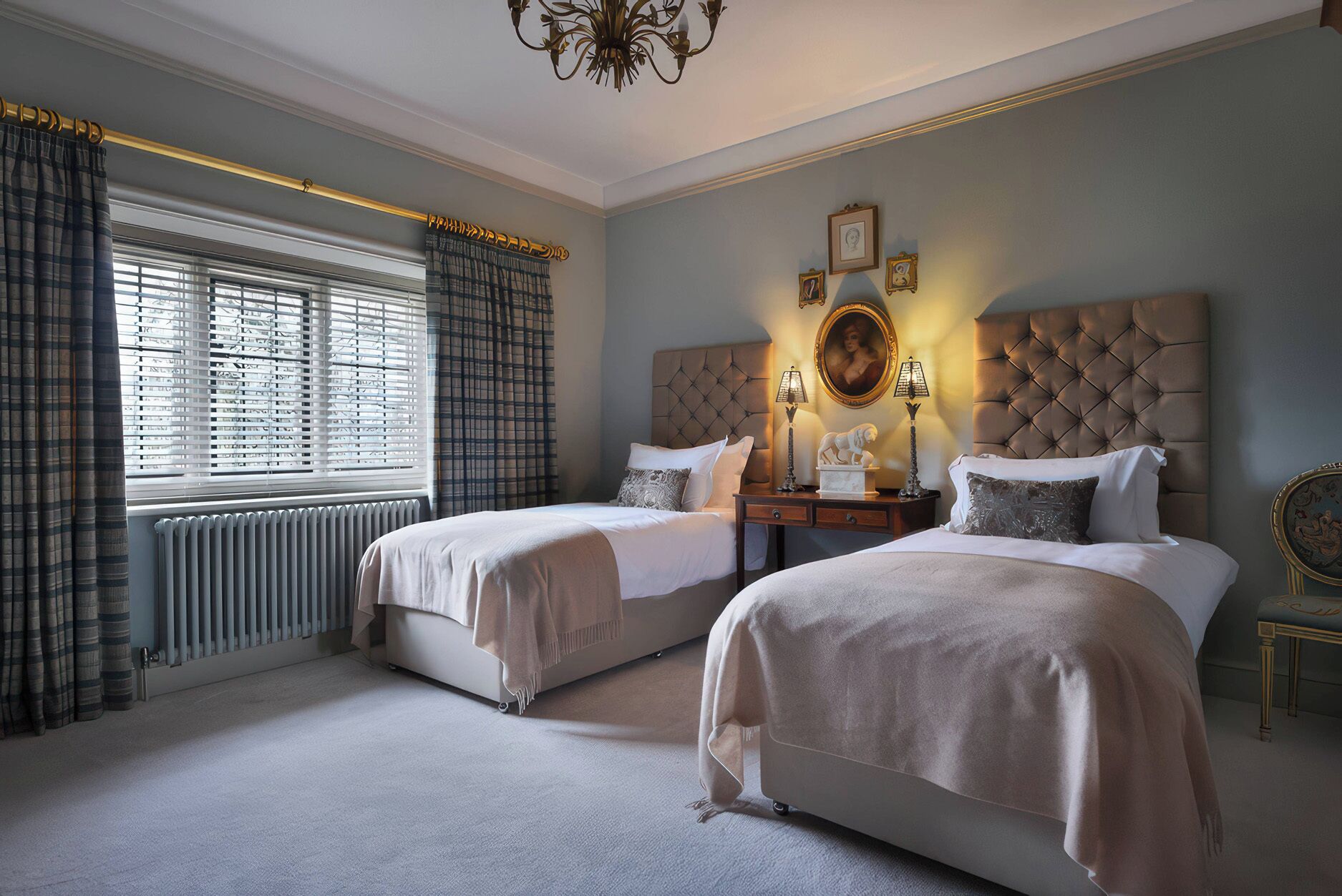 A bedroom with two beds, a radiator, a window with curtains, a chandelier, and a wall with framed pictures.