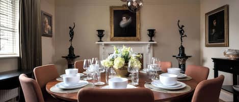 A formal dining room with a round table set for six, flanked by two black statues, a fireplace, and framed portraits on the walls.