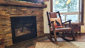 A stone fireplace, a rocking chair with a wolf pillow, and a window with a view of trees. - Hot tub, mtn views, massage chair, & steam shower! (Divide)