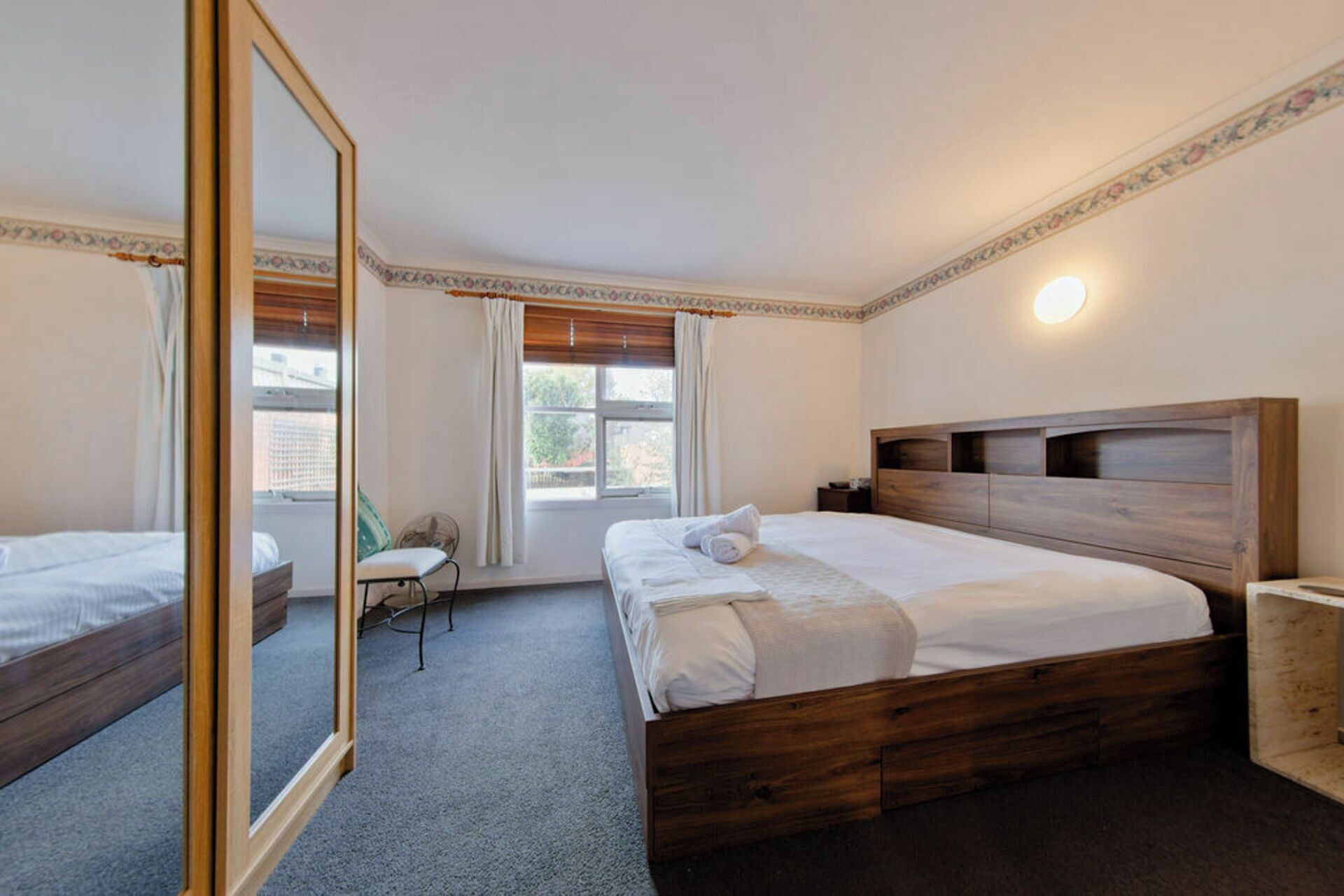 Ein Schlafzimmer mit großem Spiegel, einem Bett mit Holz-Kopfteil, einem Sessel und einem Fenster mit Vorhängen.