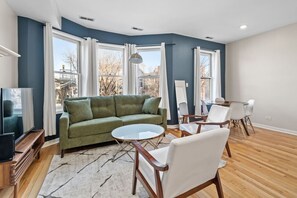 Ein modernes Wohnzimmer mit einem grünen Sofa, weißen Sesseln, einem kleinen runden Tisch und großen Fenstern.