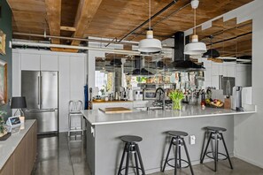 A modern kitchen with stainless steel appliances, a central island with stools, hanging pendant lights, and exposed wooden beams.