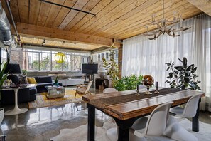 A spacious living area with a wooden ceiling, a dining table, modern furniture, and large windows.