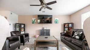 Uma sala de estar com ventilador de teto, televisão de tela plana, sofás de couro, mesa de centro e um aparador com objetos decorativos.