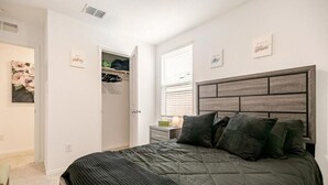 Um quarto com cama, mesa de cabeceira, janela com persianas e armário com roupas penduradas.