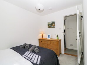 A bedroom with a bed, a wooden dresser, a nightstand with a lamp, a painting on the wall, and a door leading to another room. - Fellfoot Cottage (Cockermouth)