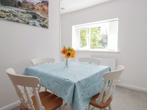 Ein Essbereich mit einem Tisch, bedeckt mit einer hellblauen Tischdecke mit Vogelmuster, umgeben von weißen Stühlen. Ein Fenster mit weißen Jalousien und ein gerahmtes Bild an der Wand sind ebenfalls zu sehen.