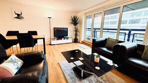 Una sala de estar moderna con un sofá de cuero negro, una mesa de centro de vidrio, un televisor de pantalla plana y un comedor con una mesa de madera y sillas negras.