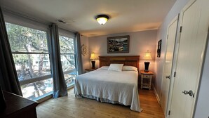 A bedroom with a bed, two bedside tables with lamps, a window with curtains, a painting on the wall, and a door. - Beautiful 2-bedroom, 2 bathroom villa in South Forest Beach. Hilton Head Island (Hilton Head Island)
