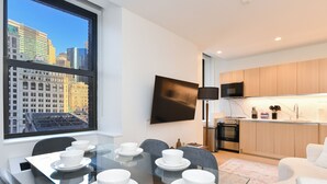 A modern kitchen with a flat-screen TV, a dining table with white bowls, and a city view through a large window. - Sleek 2BR Oasis in NYC Financial District (New York)