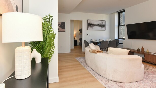 A modern living room with a sofa, a flat-screen TV, and a dining area in the background. - Sleek 2BR Oasis in NYC Financial District (New York)
