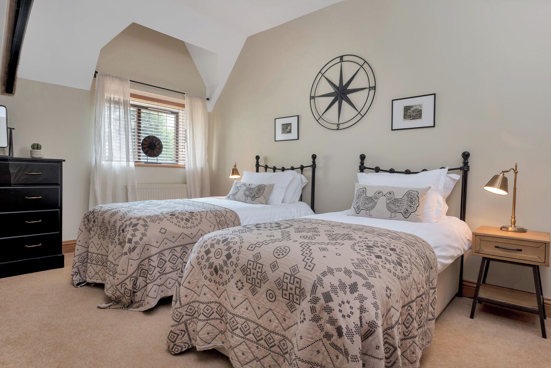 A bedroom with two beds, a dresser, a nightstand, a lamp, a compass wall decoration, and a window with curtains.