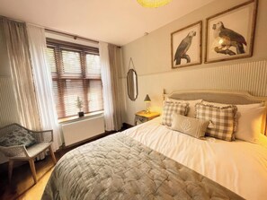 A bedroom with a bed, a chair, a window with blinds, and two framed pictures on the wall. - The Lady Rothschild Stables (Peterborough)