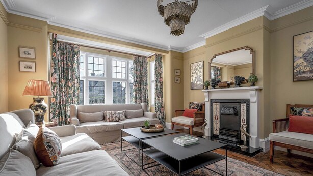 Ein traditionell eingerichtetes Wohnzimmer mit Sofa, Sesseln, Kamin und einem großen Fenster mit Vorhängen.