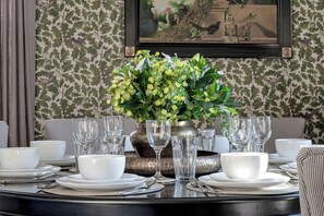 A dining table set with white plates, bowls, and clear glasses, surrounded by a patterned wallpaper and a framed painting. - Greenwood Manor (Peterborough)