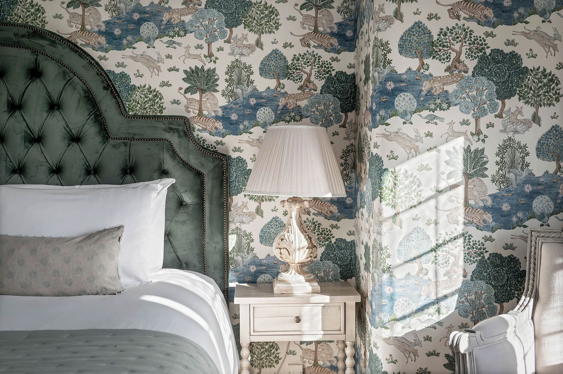 A bedroom with a tufted headboard, a bedside table with a lamp, and a window with patterned curtains.