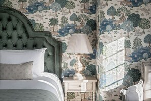 A bedroom with a tufted headboard, a bedside table with a lamp, and a window with patterned curtains. - Greenwood Manor (Peterborough)