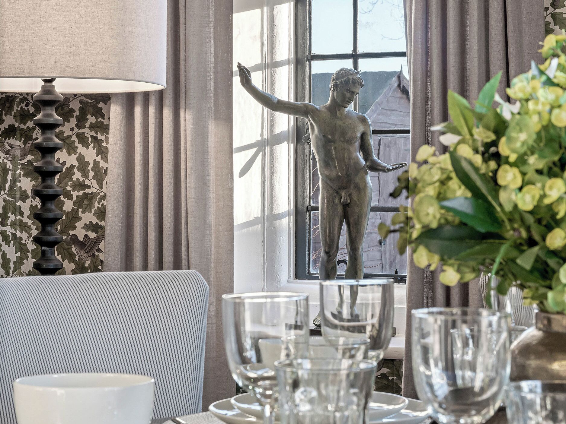 Un coin repas avec une table dressée pour deux personnes, une statue et une fenêtre avec des rideaux.
