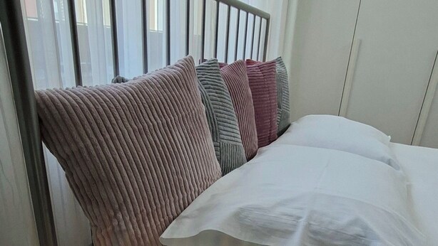 Una cama con ropa de cama blanca, un cabecero de metal y varias almohadas. Hay una mesita de cristal con una lámpara junto a la cama.