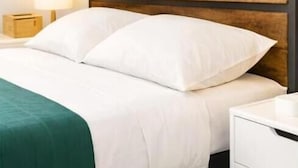 A bed with white bedding, a wooden headboard, a green throw blanket, a bedside lamp, and a white nightstand.