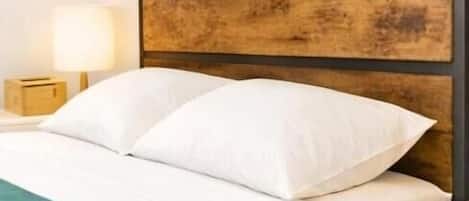 Una cama con ropa de cama blanca, cabecera de madera, manta verde y una lámpara de noche. También se observa una mesita de noche blanca.