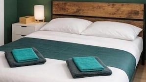 A bed with white linens, a teal bedspread, and two teal towels. There is a wooden headboard, a nightstand with a lamp, and two framed pictures on the wall.