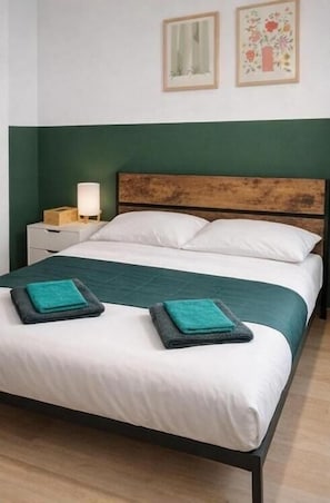 Ein Bett mit weißen Leinentüchern, einer türkisblauen Tagesdecke und zwei türkisblauen Handtüchern. Dazu ein hölzerner Kopfteil, ein Nachttisch mit Lampe sowie zwei gerahmte Bilder an der Wand.