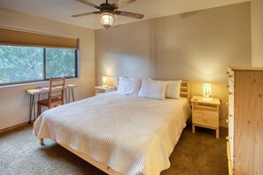 A bedroom with a large bed, a desk, and a chair, a ceiling fan, and a window with a view of trees. - Camp Klip at Bison Ridge (Show Low)
