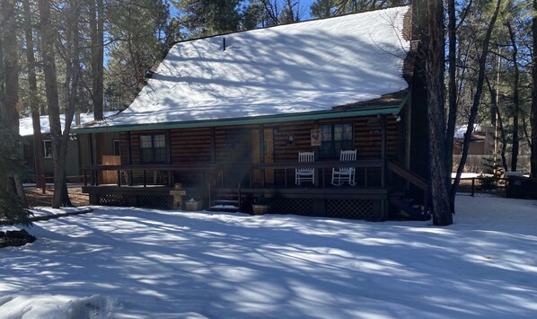 Une cabane en rondins avec un porche, entourée de neige et d’arbres.