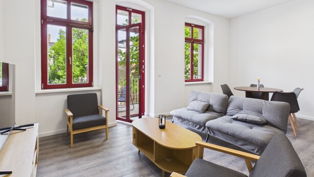 A modern living room with a grey sofa, wooden coffee table, and two armchairs. Large windows with red frames offer views of greenery outside. - Group apartment with balcony, up to 5 persons - GÖ05 (Görlitz)