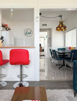 Une cuisine moderne avec une pancarte indiquant « Ici, on sert de bons plats », un coin repas avec une table ronde et des chaises, et un bar avec des chaises rouges.