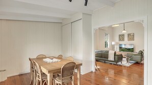 Un comedor con una mesa de madera y sillas de mimbre, una sala de estar con sofá y televisor, y un pasillo con paredes blancas y pisos de madera.