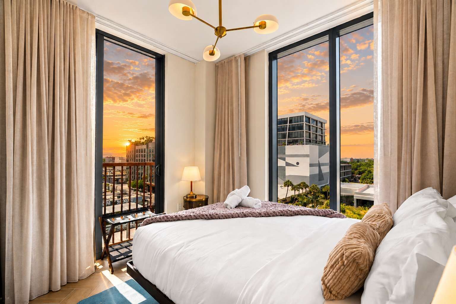 Quarto de hotel com uma cama grande, uma mesa de cabeceira, um abajur e vista para o pôr do sol sobre a paisagem urbana.