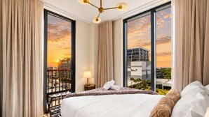 A hotel room with a large bed, a nightstand, a lamp, and a view of a sunset over a cityscape. - Modern 2BR | Pool | Steps to Miami's Best Murals (Miami)