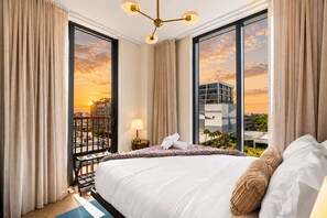 Ein Hotelzimmer mit einem großen Bett, einem Nachttisch, einer Lampe und Blick auf einen Sonnenuntergang über einer Stadtlandschaft.