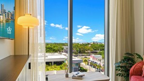 A modern room with a large window, a desk with a lamp, a red sofa, and a view of a cityscape. - Modern 2BR | Pool | Steps to Miami's Best Murals (Miami)