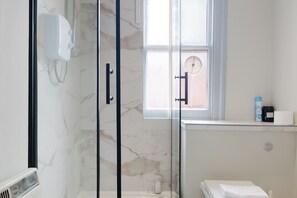 Ein modernes Badezimmer mit Duschkabine aus Glas, Wandverkleidung aus weißem Marmor, Fenster mit Handtuchhalter und eine kleine Ablage für Toilettenartikel.