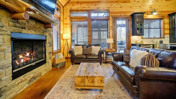 A log cabin interior with a stone fireplace, leather sofas, a wooden coffee table, and a kitchen area. - Secluded Cabin, Hot Tub, Game Loft, 2 Fireplaces (Broken Bow)