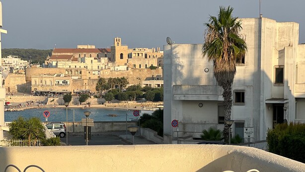 Un balcón con dos sillas blancas de metal y una mesa, con vistas a un pueblo costero con edificios y una palmera.