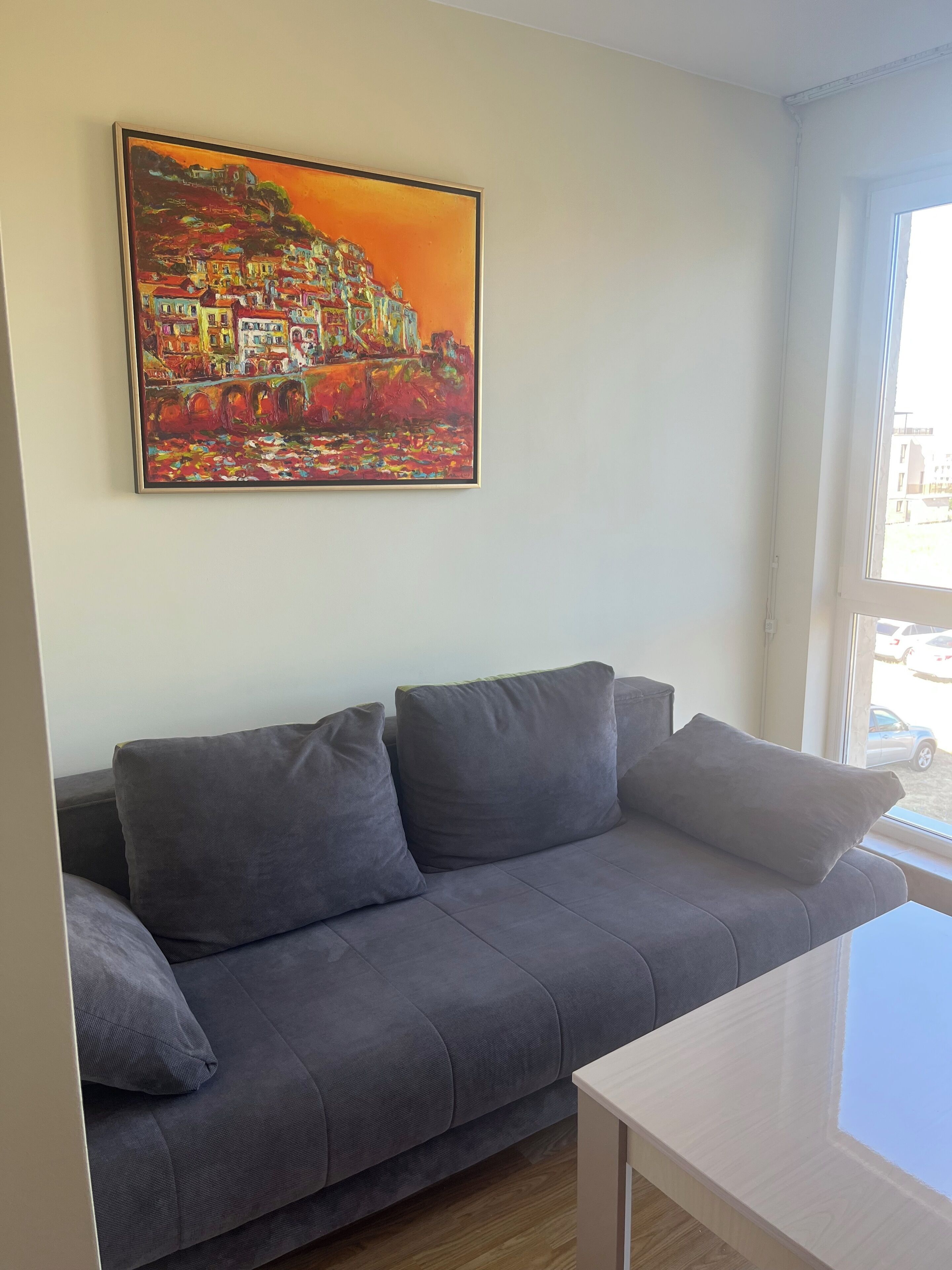 A room with a grey sofa, a painting of a colorful landscape, and a window with a view of the outside.