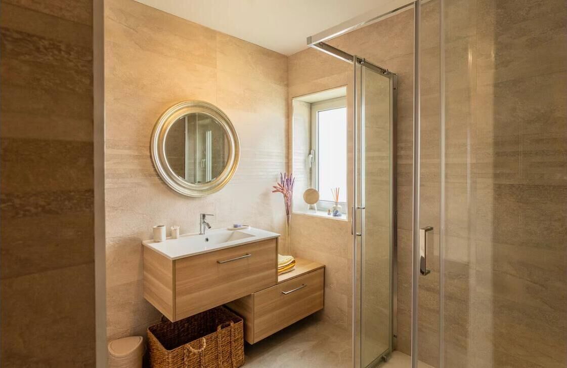 A modern bathroom with a glass shower enclosure, a round mirror, and a wooden vanity.
