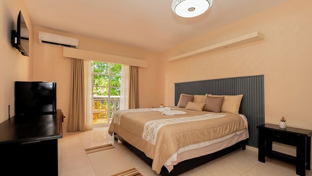 Une chambre à coucher avec un lit, une télévision, une table de chevet, un luminaire au plafond et une fenêtre avec des rideaux.